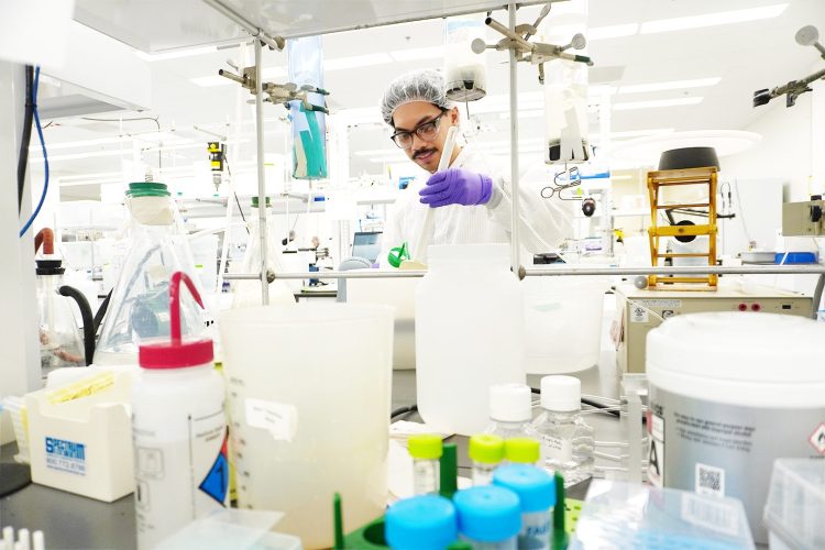 A laboratory technician wearing safety goggles, a hairnet, and purple gloves works with lab equipment in a bright, sterile laboratory environment. Various containers, beakers, test tubes, and scientific instruments surround the workspace, indicating active scientific research or experimentation.