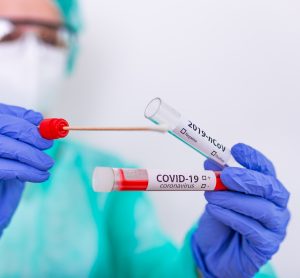 Nurse in gown, face mask and hloves holding a nasal swab and inserting it into a tube labelled 'COVID-19 Test'