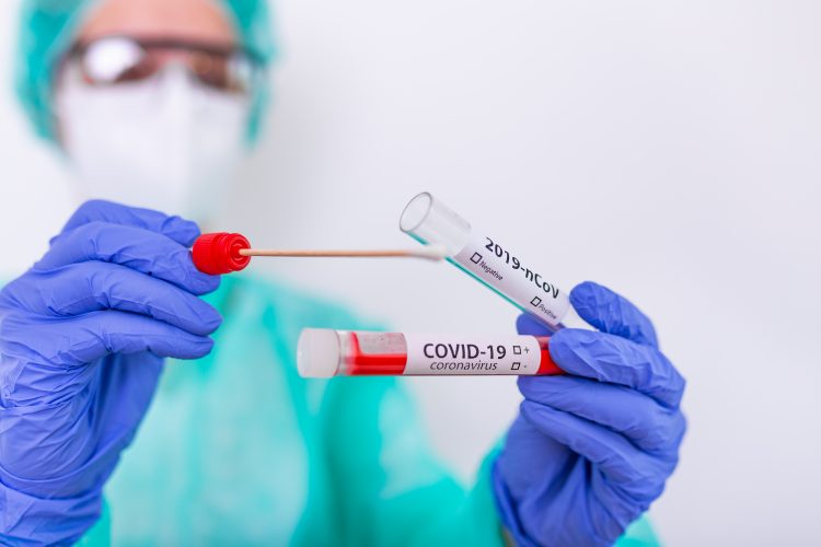 Nurse in gown, face mask and hloves holding a nasal swab and inserting it into a tube labelled 'COVID-19 Test'