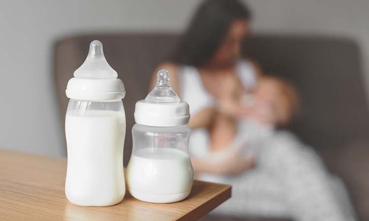 Bottles with breast milk on the background of mother holding in her hands and breastfeeding baby. Maternity and baby care.
