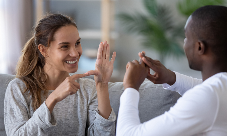 Smiling interracial friends talking with sign finger hand language, happy two deaf and mute hearing impaired people communicating at home sit on sofa showing hand gestures