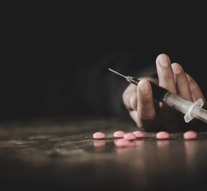 A drug addict reaches for another dose of the heroin in the syringe. Hand surrounded by pink pills.