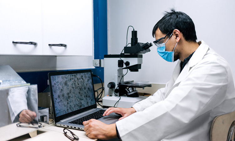 Stock photo of scientist wearing face mask using the computer and a microscopy in his lab.