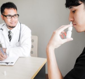 man using a nasal spray being observed by a researcher/doctor in a lab coat, making notes on a clipboard