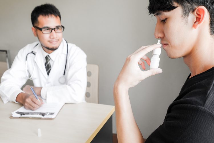 man using a nasal spray being observed by a researcher/doctor in a lab coat, making notes on a clipboard