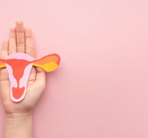 Hand holding uterus shape made from paper on pink background.