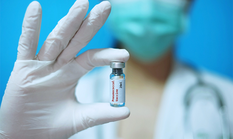 A physician with surgical mask and white rubber gloves at a clinic, holding a glass bottle of 1 dose of pneumococcal disease vaccine with white background and red letters.