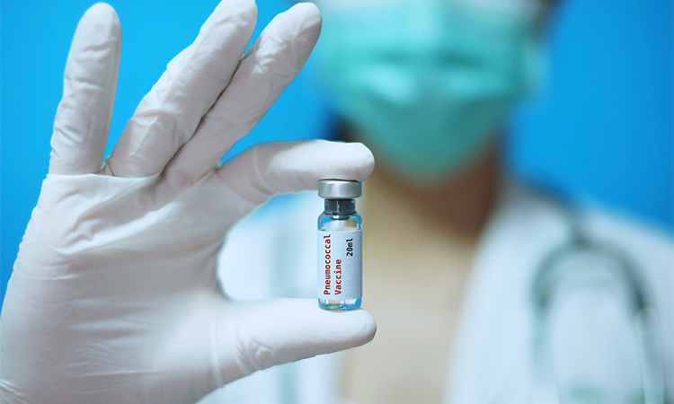 A physician with surgical mask and white rubber gloves at a clinic, holding a glass bottle of 1 dose of pneumococcal disease vaccine with white background and red letters.