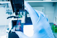 A gloved hand holding a next-generation sequencing flow cell in a bright laboratory setting with scientific equipment in the background.