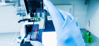 A gloved hand holding a next-generation sequencing flow cell in a bright laboratory setting with scientific equipment in the background.