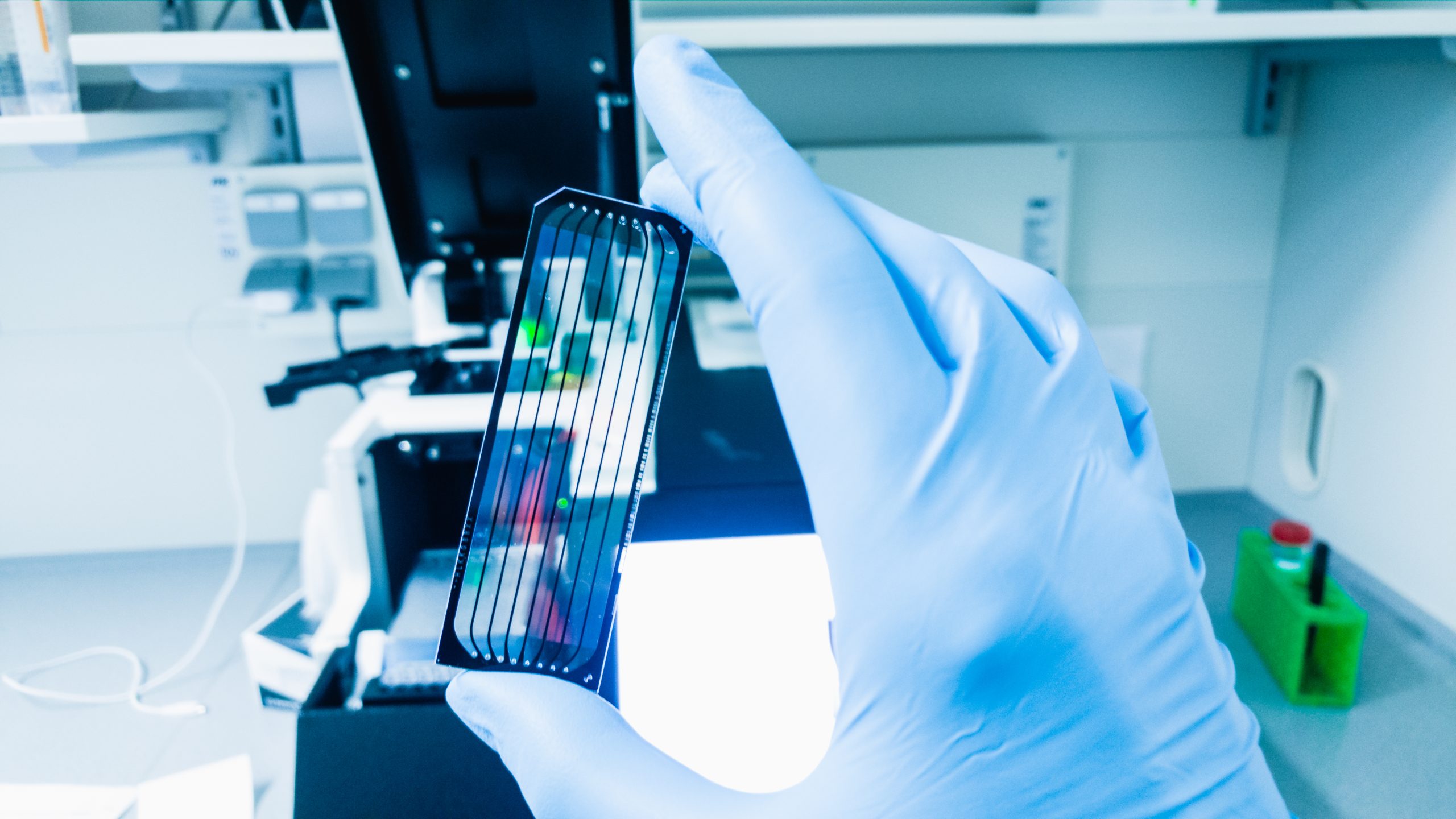 A gloved hand holding a next-generation sequencing flow cell in a bright laboratory setting with scientific equipment in the background.