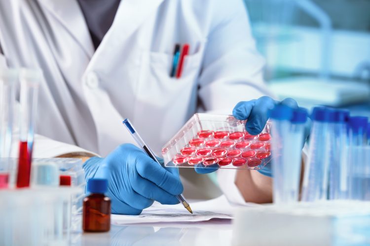 Scientist in a lab coat and gloves analysing samples in a multiwell plate, surrounded by test tubes and lab equipment