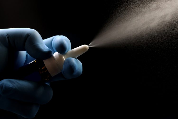 A gloved hand spraying a nasal spray, showing a fine mist against a dark background, representing innovation in nasal drug delivery technology.
