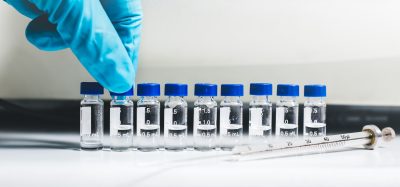 Gloved hand arranging a row of small clear laboratory vials with blue caps, partially filled with liquid, next to a glass syringe on a white lab bench.