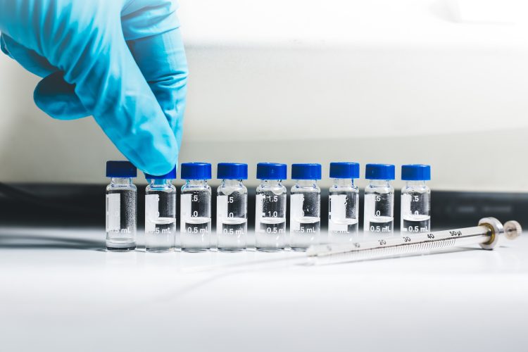 Gloved hand arranging a row of small clear laboratory vials with blue caps, partially filled with liquid, next to a glass syringe on a white lab bench.