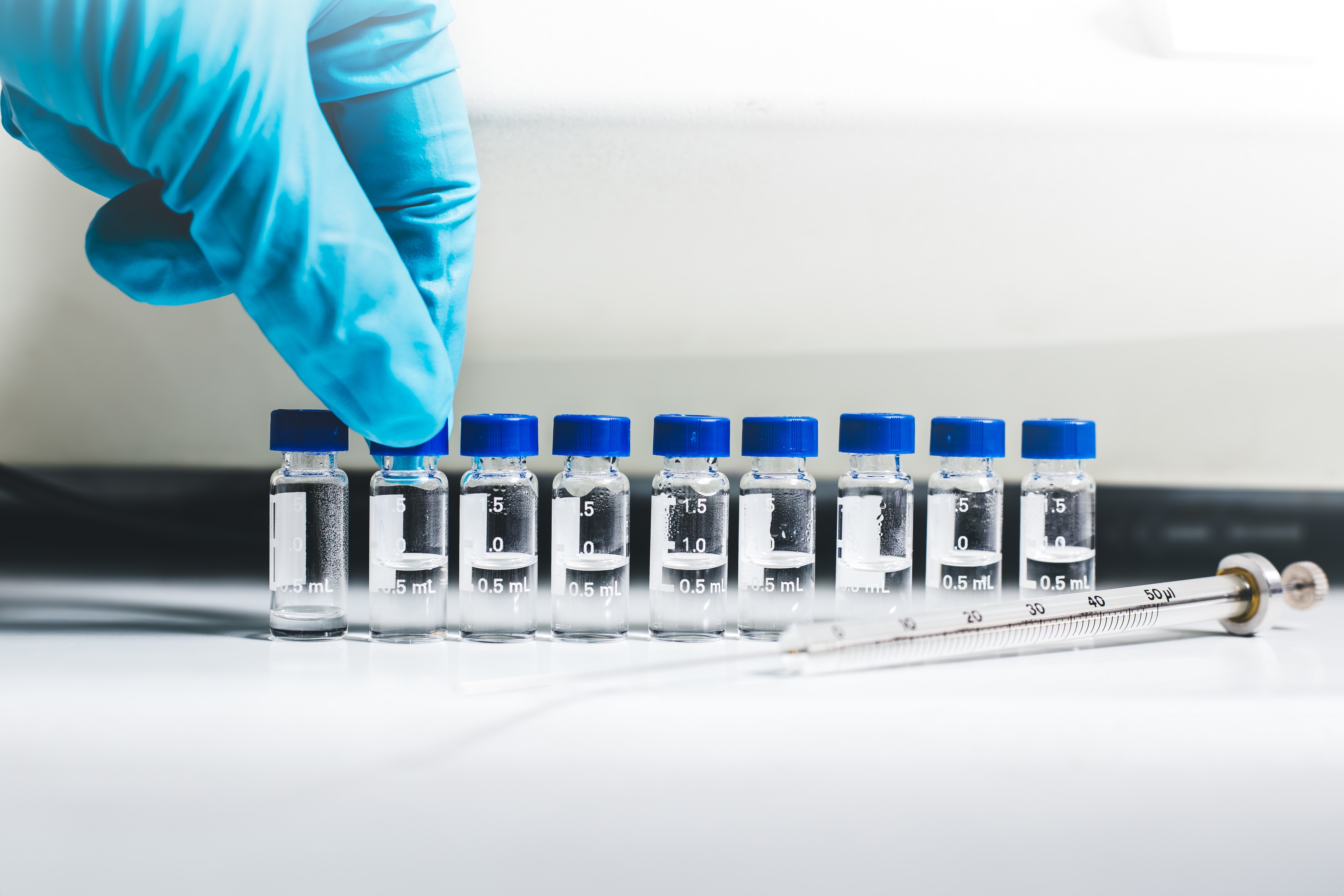 Gloved hand arranging a row of small clear laboratory vials with blue caps, partially filled with liquid, next to a glass syringe on a white lab bench.