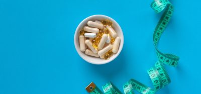 A white bowl filled with capsules and softgel pills next to a green measuring tape on a blue background, symbolising medical treatments and weight management in obesity care.