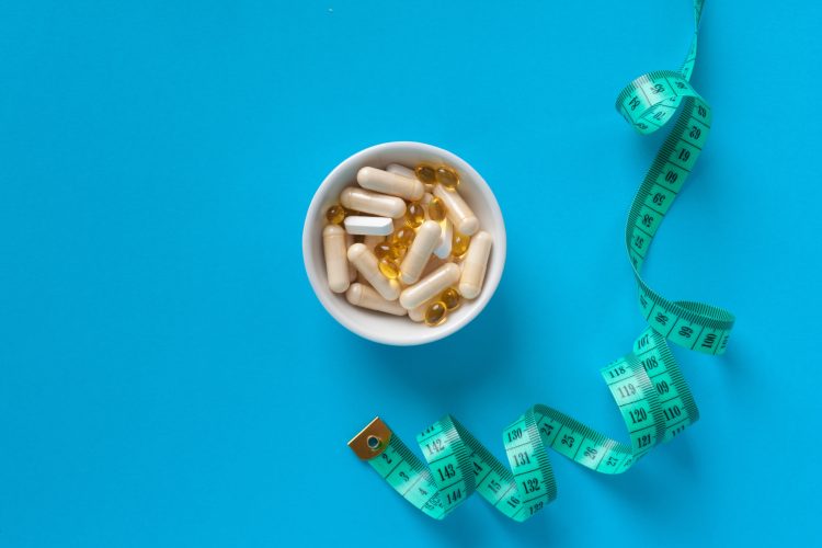 A white bowl filled with capsules and softgel pills next to a green measuring tape on a blue background, symbolising medical treatments and weight management in obesity care.