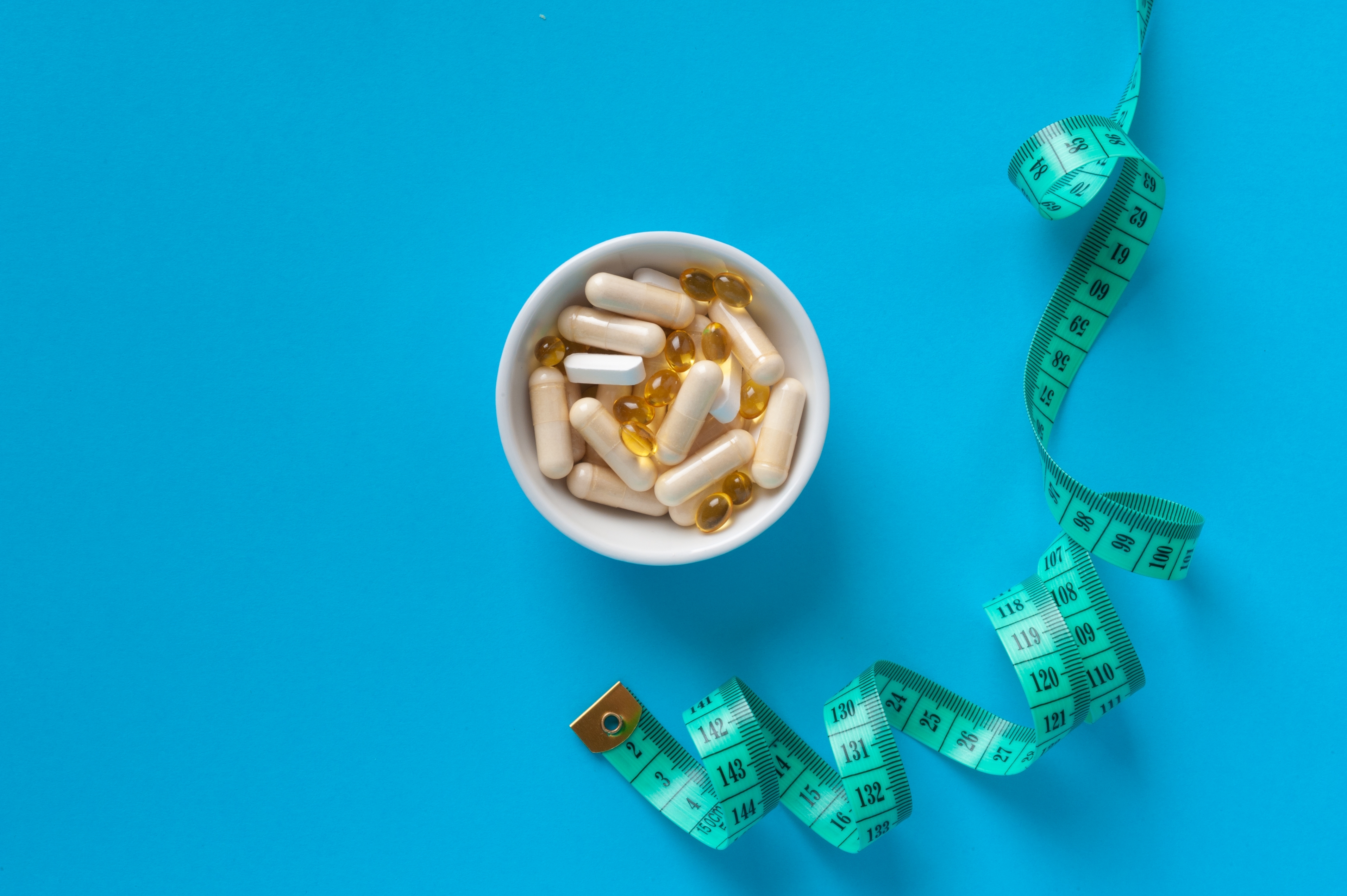 A white bowl filled with capsules and softgel pills next to a green measuring tape on a blue background, symbolising medical treatments and weight management in obesity care.