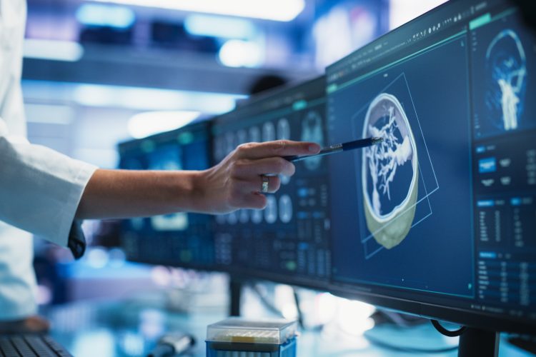 Researcher in a lab coat points to a brain scan on a computer monitor, highlighting areas of interest for analysis of brain tumours.