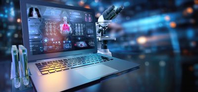 A laptop displaying a digital biomedical dashboard sits beside a laboratory microscope and test tubes, representing the integration of AI, data analysis, and laboratory research.