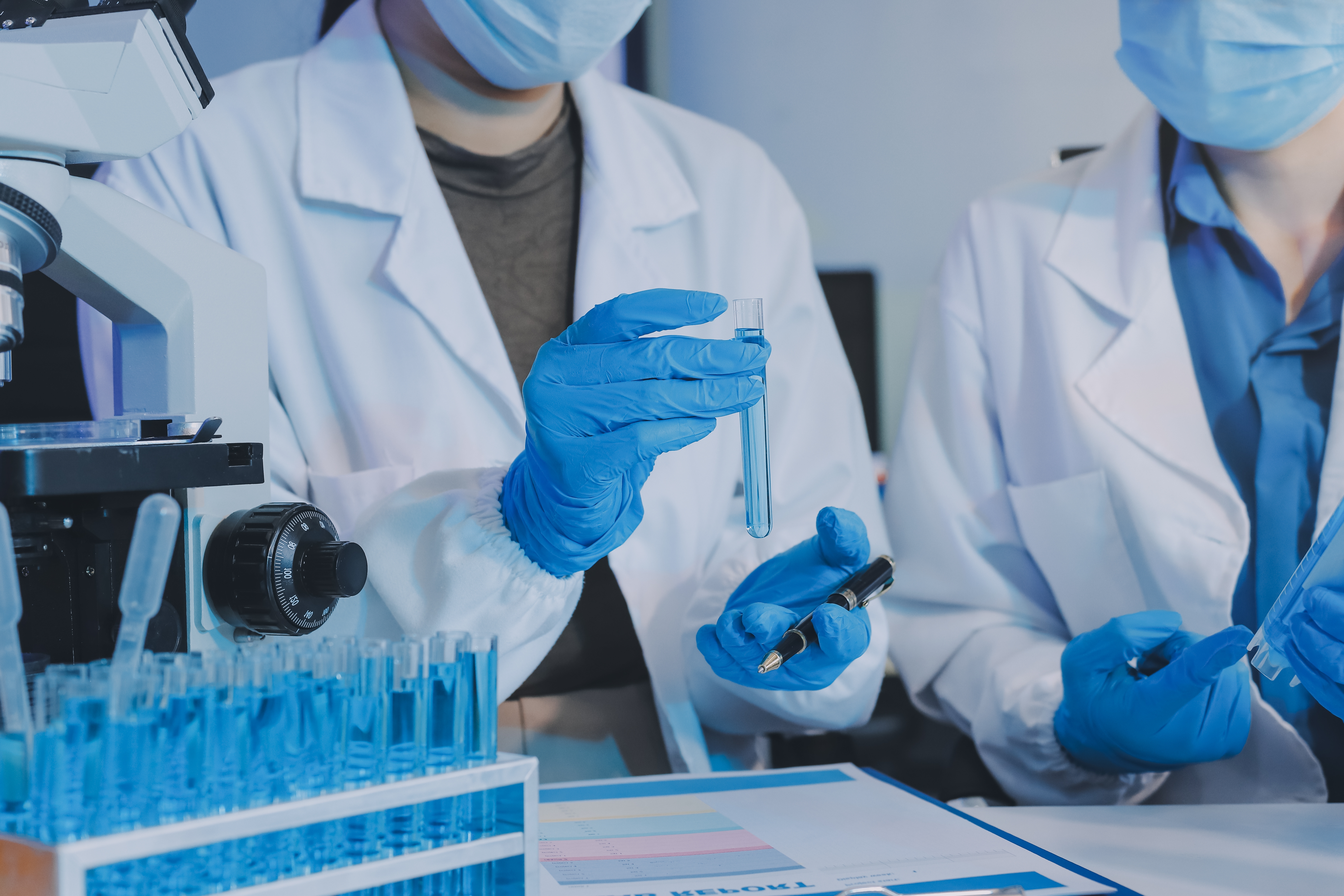 Scientists in a laboratory examining test tubes with blue liquid beside a microscope.